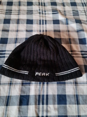 Svart ribbstickad mössa Peak - Svart ribbstickad mössa från Peak med vita ränder och broderad logga framtill. Klassisk modell som sitter skönt på huvudet och passar perfekt när det är kallt ute. Enkel och stilren design som funkar till det mesta.