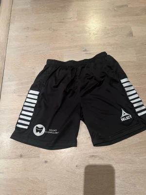 Svarta sportshorts från Select, stl 164/14y - Svarta sportshorts från Select med vita ränder på sidorna och tryck av Malmö Andrelag. Shorts i lätt och snabbtorkande material med resår i midjan, perfekta för träning eller match⚽️🤾‍♂️