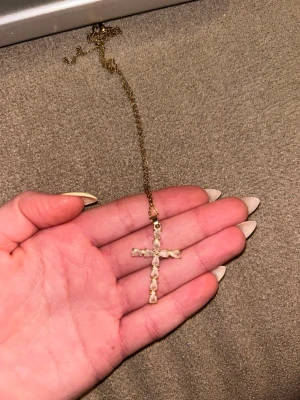 Guldfärgat halsband med korshänge - Snyggt halsband i guldfärg med ett korsformat hänge dekorerat med små vita stenar. Kedjan är tunn och har ett klassiskt lås. Perfekt accessoar för dig som gillar stilrena smycken med lite bling.