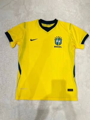 Gul Brasil Nike fotbollströja - Gul Brasilien fotbollströja från Nike med gröna detaljer och CBF-emblem på bröstet. Tröjan har korta ärmar, V-ringad krage och är tillverkad i Dri-FIT material som andas. Perfekt för fotbollsträning eller att bära när du hejar på landslaget.