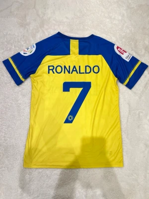 Al Nassr Ronaldo matchtröja gul/blå - Säljer en Al Nassr matchtröja i gult och blått med Ronaldo och nummer 7 på ryggen. Tröjan har klubbmärke, sponsorloggor och är tillverkad i lätt, ventilerande material perfekt för fotbollsträning eller match.