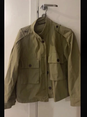 Oliv utilityjacka med fickor - Stilren oliv utilityjacka med stora fickor framtill och axelremmar. Jackan har ståkrage, knappar och lång ärm. Perfekt för dig som gillar en avslappnad och trendig look.