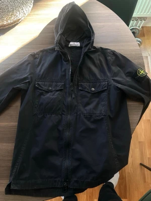 Svart vindjacka från Stone Island - Svart vindjacka från Stone Island med huva och dragkedja framtill. Jackan har två bröstfickor med knappar och den klassiska Stone Island-loggan på ärmen. Tillverkad i bomull. Storlek S. Sparsamt använd på senare år, observera att de är lite missfärgningar på axlarna men inget som syns eller påverkar jackan. 