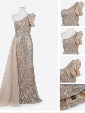 Glittrig långklänning med blomdetalj - En elegant långklänning i champagne och silver med glittrigt mönster, oneshoulder och stor blomdetalj på axeln. Klänningen har en lång, flytande chiffongpanel på sidan och figurnära passform. Perfekt för bal eller festliga tillfällen.