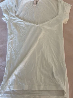 Vit v-ringad topp från Nelly  - Vit kortärmad t-shirt med spetskant vid halsen. Modellen är figurnära och har en klassisk passform. Den är ifrån Nelly i storlek xxs