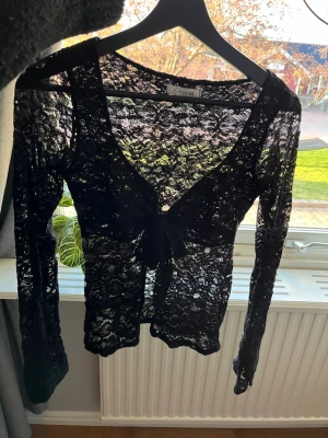Svart spetsig knytblus med lång ärm - Snygg svart spetsblus från Design by Si med v-ringning och knyt framtill. Blusen är genombruten i spetsmaterial och har långa ärmar. Perfekt att styla över ett linne eller topp för en trendig look.