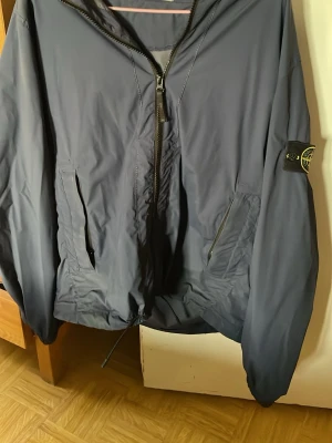Mörkblå vindjacka från Stone Island - Snygg mörkblå vindjacka från Stone Island med dragkedja framtill och två fickor med dragkedja. Jackan har klassisk logotyp-patch på ärmen och justerbar nederkant. Perfekt för blåsiga dagar och streetwear-stil.