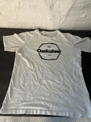 Vit Quiksilver t-shirt med tryck - Vit t-shirt från Quiksilver med ett svart tryck och logga framtill. Klassisk passform med rund hals och korta ärmar. Tillverkad i mjuk bomull, perfekt för en avslappnad stil.