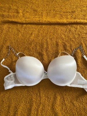 Vit push-up bh med kedjedetaljer - Vit push-up bh med vadderade kupor och unika axelband i guldfärgad kedja. Bh:n har släta kupor och justerbara band. Perfekt för dig som vill ha något extra till outfiten.