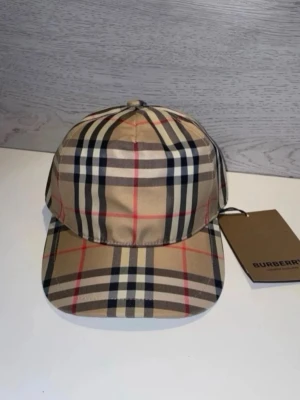 Beige keps från Burberry med rutmönster - Säljer en klassisk beige keps från Burberry med det ikoniska rutmönstret i svart, vitt och rött. Kepsen har böjd skärm och justerbar rem bak. Tillverkad i ett mjukt och slitstarkt material som ger en lyxig känsla.