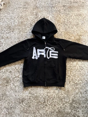 Svart zip hoodie från Arte - Säljer en svart hoodie med dragkedja från Arte. Stor vit logga i graffiti-stil på bröstet och klassisk huva. Hoodien har känguruficka framtill och ribbade muddar vid ärmar och nederkant. Perfekt för en chill och streetwear-inspirerad look.