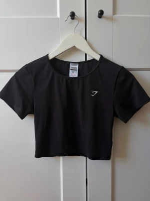 Svart croppad topp från Gymshark - Snygg svart croppad topp från Gymshark med rund halsringning och korta ärmar. Toppen har en diskret logga på bröstet och är tillverkad i ett stretchigt syntetmaterial som passar perfekt till träning.