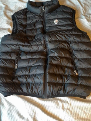 Svart dunväst från Moncler - Snygg svart dunväst från Moncler med klassisk quiltad design och hög krage. Västen har två dragkedjeförsedda fickor framtill och Moncler-logga på bröstet. Perfekt för lager-på-lager och håller dig varm med stil.