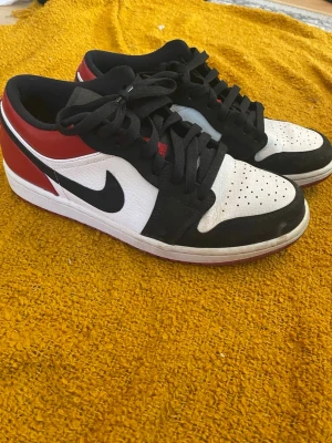 Nike Air Jordan 1 Low svart/vit/röd - Snygga Nike Air Jordan 1 Low sneakers i svart, vitt och rött med klassisk Swoosh-logga på sidan. Ovandelen är i läder med perforerad tåbox och svarta skosnören. Yttersulan är röd och har bra grepp. Perfekta för dig som gillar streetwear och ikoniska sneakers.