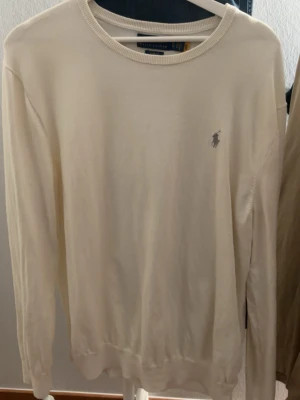 Polo Ralph Lauren tröja  - Klassisk cremevit tröja från Polo Ralph Lauren med rund halsringning och diskret broderad logga på bröstet. Tröjan har lång ärm och är tillverkad i mjukt material som känns skönt mot huden. Perfekt för en stilren look.