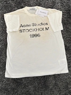 Vit Acne Studios t-shirt med tryck - Vit t-shirt från Acne Studios med svart texttryck 'Acne Studios STOCKHOLM 1996' på bröstet. Klassisk rund hals och korta ärmar. Tillverkad i mjuk bomull, perfekt för en stilren och avslappnad look.