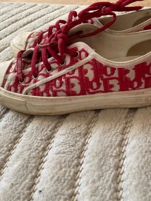 Röda Dior sneakers - Snygga låga sneakers från Dior med det klassiska Oblique-mönstret i rött och vitt. Skorna har röda snören, vit sula och detaljer i canvasmaterial. Perfekta för dig som vill sticka ut med en ikonisk designerstil.