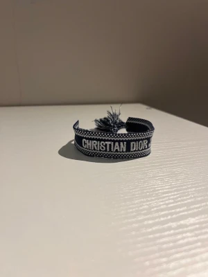 Christian Dior J'adior tygarmband - Snyggt marinblått tygarmband från Christian Dior med texten 'CHRISTIAN DIOR J'ADIOR' broderad i vitt. Armbandet har knytning med tofsar och en cool, ungdomlig vibe. Perfekt accessoar för att lyfta din outfit.