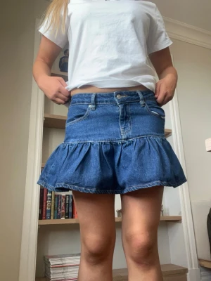Blå volang jeanskjol - Säljer en blå jeanskjol med volangkant och klassiska fem fickor. Kjolen har en kort, utställd modell och stängs med knapp och dragkedja framtill. Tillverkad i denim med synliga sömmar för en trendig look.