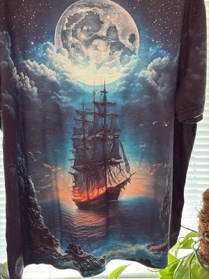 Svart t-shirt med skepp och måne storlek 40 - Unik svart t-shirt med ett stort tryck av ett seglande skepp i solnedgång under en gigantisk måne och stjärnklar himmel. Färgerna går i blått, orange och vitt, vilket ger en riktigt cool och mystisk vibe. Perfekt för dig som gillar statement-plagg och snygga motiv.