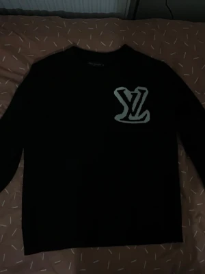 Svart stickad tröja Louis Vuitton - Snygg svart stickad tröja från Louis Vuitton med stort vitt LV-logotyp på bröstet. Tröjan har rund halsringning och långa ärmar. Perfekt för dig som gillar exklusiva märken och vill ha en stilren men ändå iögonfallande look.