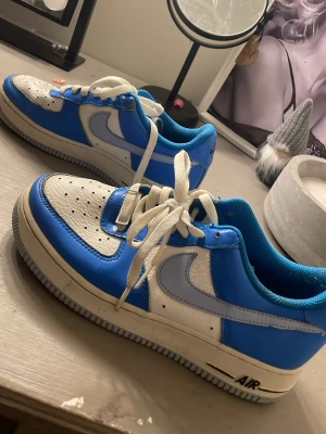 Nike Air Force 1 blå och vita sneakers - Säljer ett par Nike Air Force 1 sneakers i blått och vitt med klassisk siluett. Skorna har vit tåbox med perforeringar, blåa paneler och vit swoosh på sidorna. Yttermaterialet är läder och de har vita skosnören samt blå insida. Perfekta för en streetwear-look.