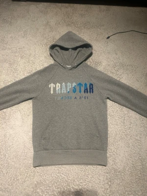 Grå Trapstar hoodie med tryck - Säljer en grå hoodie från Trapstar med huva och trycket 'TRAPSTAR IT'S A SECRET' i vitt och blått på bröstet. Hoodien har ribbade muddar och en klassisk passform. Perfekt för dig som gillar streetwear och vill ha något stilrent men ändå med attityd.