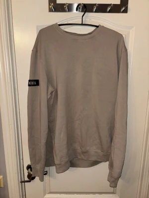Beige sweatshirt med REBEL-tryck - En stilren beige sweatshirt med rund hals och långa ärmar. På vänster ärm finns ett svart tygmärke med texten 'REBEL'. Tröjan har ribbade muddar vid ärmslut och nederkant. Perfekt för en avslappnad och trendig look.