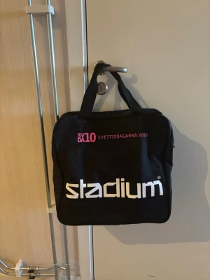 Svart duffelväska Stadium - Säljer en svart duffelväska/sportväska från Stadium med tryck i vitt och rosa. Väskan har två korta handtag, fyrkantig form och dragkedja upptill. På ena sidan står det 'stadium' och på andra 'SXD10 SVETTISDAGARNA 2010'. Perfekt för gymmet eller skolan