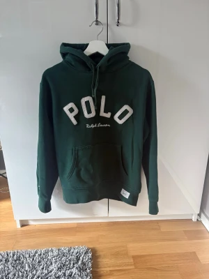 Mörkgrön hoodie från Polo Ralph Lauren - Mörkgrön hoodie med stor POLO-text i vitt på bröstet och broderad Ralph Lauren-logga under. Klassisk känguruficka framtill, huva och ribbade muddar. Perfekt för en avslappnad och stilren look.