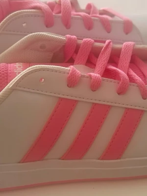 Vita och rosa sneakers med ränder - Snygga sneakers i vitt med rosa detaljer och klassiska tre ränder på sidan. Skorna har rosa skosnören och en låg siluett. Ovandelen är i syntetmaterial och insidan är ljusbeige. Perfekta för dig som gillar färg och sportig stil.