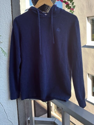 Merinoull hoodie - En stilren mörkblå hoodie med huva och dragsnören. Tröjan har lång ärm och ett diskret broderat märke på bröstet. Tillverkad i mjukt stickat material som känns skönt mot huden. Perfekt för en avslappnad look. Använd fåtal gånger och pris ej hugget i sten. Nypris: 1199kr