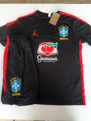 Svart Brasilien träningsset Jordan - Snyggt svart träningsset med Brasilien-tryck från Jordan. Setet består av en t-shirt och shorts med röda detaljer längs sidorna, CBF-logga och Guaraná Antarctica sponsortryck. Materialet är lätt och andas, perfekt för fotbollsträning eller gymmet.