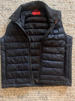 Hugo Boss - mörkblå dunväst med dragkedja - Snygg mörkblå dunväst med quiltad design, röda detaljer och hög krage från Hugo Boss. Västen har en praktisk dragkedja framtill och är perfekt att slänga över en hoodie eller tröja för extra värme. 