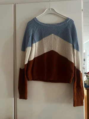Stickad tröja med färgblock i blått - Säljer en stickad tröja med breda färgblock i ljusblått, vitt och rostbrunt. Tröjan har rund halsringning, lång ärm och ribbstickade muddar. Perfekt för dig som gillar retrovibbar och vill ha något mysigt och trendigt i garderoben.