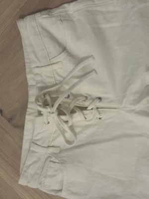Vita jeans med snörning fram - Säljer ett par vita jeans med unik snörning framtill istället för dragkedja. Byxorna har raka ben och klassiska fickor fram och bak. Materialet ser ut att vara bomull med lite stretch för extra komfort. Perfekta för dig som vill sticka ut med detaljer!