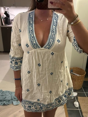 Vit och blå kortklänning med v-ringning - Säljer en luftig vit kortklänning med blå broderade detaljer och v-ringning. Klänningen har trekvartsärm och ett blommigt mönster samt dekorativa kanter. Perfekt för sommarens varma dagar och har en avslappnad passform.