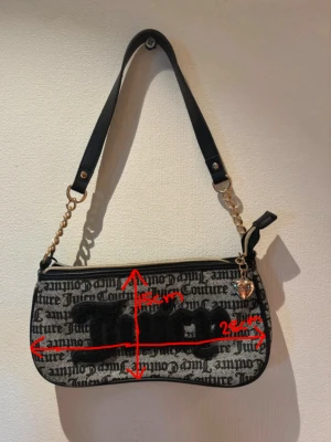 Juicy Couture svart axelväska - Juicy Couture axelväska i svart och grått med tydligt logomönster och stor svart 'Juicy'-text framtill. Väskan har guldfärgade kedjedetaljer, svart axelrem och ett hjärtformat hänge vid dragkedjan. Materialet är textil med detaljer i konstläder.