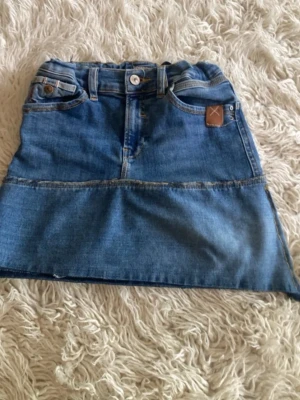 Blå jeanskjol från LTB - Snygg blå jeanskjol från LTB med klassisk femficksdesign och knapp- samt dragkedjestängning. Kjolen har en rak passform och är sydd i denim med synliga sömmar och läderpatch bak i midjan. 