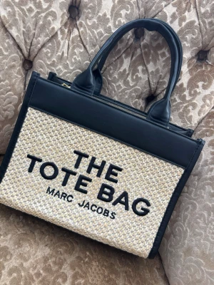 A The Tote Bag utan axelrem  - Snygg handväska  i modellen The Tote Bag. Väskan har svarta handtag och detaljer i läder samt beige flätad front med tydlig logga. 20 cm hög och 27 cm bred . Oandvänd 