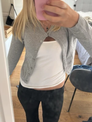 Grå cropped kofta med knappar - Säljer en ljusgrå cropped kofta med långa ärmar och ribbade muddar. Koftan har rund halsringning och stängs med flera små knappar framtill. Perfekt att slänga över ett linne eller t-shirt för en chill look.