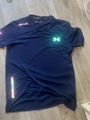 Blå tränings-tshirt från Under Armour - Snygg mörkblå tränings-tshirt från Under Armour med reflekterande logga på bröstet och ärmen. Tillverkad i lätt och snabbtorkande material, perfekt för löpning eller gym. V-ringad hals och korta ärmar ger en sportig look.