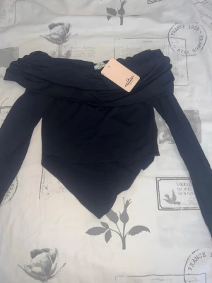 Svart offshoulder topp med lång ärm - Trendig svart offshoulder topp med långa ärmar och asymmetrisk nederkant. Toppen har en croppad passform och är perfekt för dig som vill ha en stilren och modern look. Tillverkad i mjukt material som sitter skönt på kroppen.