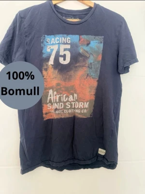 Blå t-shirt med tryck  - Snygg mörkblå t-shirt med stort färgglatt tryck på bröstet där det står 'Racing 75 African Sand Storm'. T-shirten är i 100% bomull och har en klassisk passform med rund hals och korta ärmar.
