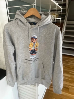 Grå hoodie Polo Bear Ralph Lauren - Snygg grå hoodie från Ralph Lauren med Polo Bear-tryck på bröstet. Klassisk passform med huva och dragsnören, samt magficka framtill. Tillverkad i mjukt bomullsmaterial som är skönt att ha på sig. Perfekt för en avslappnad och trendig look.