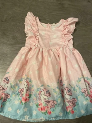Rosa enhörningsklänning med volang - Söt rosa klänning med volangdetaljer på axlarna och ett färgglatt enhörningsmönster längs nederkanten. Klänningen har vita stjärnor och blommor mot en ljusblå botten. Perfekt för dig som älskar magiska motiv!