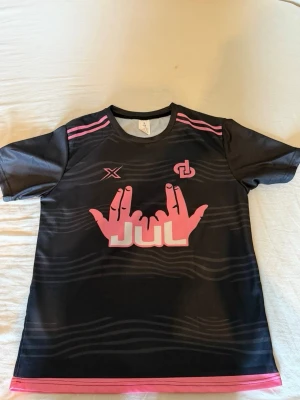Svart sporttröja med rosa tryck - Snygg svart sporttröja med rosa detaljer och tryck framtill. Tröjan har korta ärmar, rund hals och ett stort grafiskt motiv med händer och texten 'JUL' i rosa och vitt. Tillverkad i ett lätt och andningsbart material, perfekt för träning eller sport.