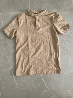 Beige randig henley med kort ärm - Snygg beige henley-skjorta med tunna vita ränder och korta ärmar. Skjortan har en klassisk knäppning med tre knappar framtill och en mjuk, bekväm känsla. Perfekt för en avslappnad och stilren look.