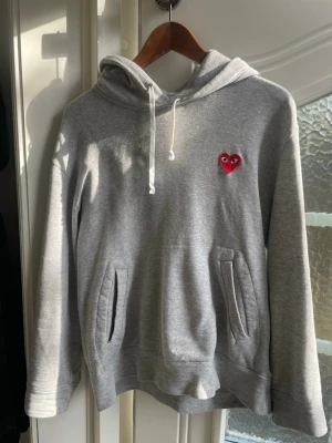 Grå Comme des Garçons hoodie - Säljer en grå hoodie från Comme des Garçons Play med det ikoniska röda hjärtat med ögon broderat på bröstet. Hoodien har huva med dragsko, magficka och ribbade muddar. Tillverkad i mjuk bomullsblandning som är riktigt skön att ha på sig.