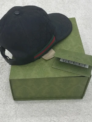Svart keps från Gucci med röd/grön rand - Snygg svart äkta keps från Gucci med klassisk röd och grön rand på sidan. Kepsen har böjd skärm och justerbart spänne bak. Tillverkad i tyg med diskret GG-mönster i materialet. Perfekt accessoar för en trendig och exklusiv look.(priset kan diskuteras)